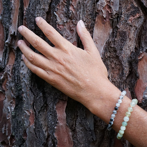 Gros plan d'une main avec des bracelets en perles posée sur l'écorce rugueuse d'un pin.